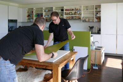 Zwei Männer platzieren grüne Folie auf Küchenschranktür.